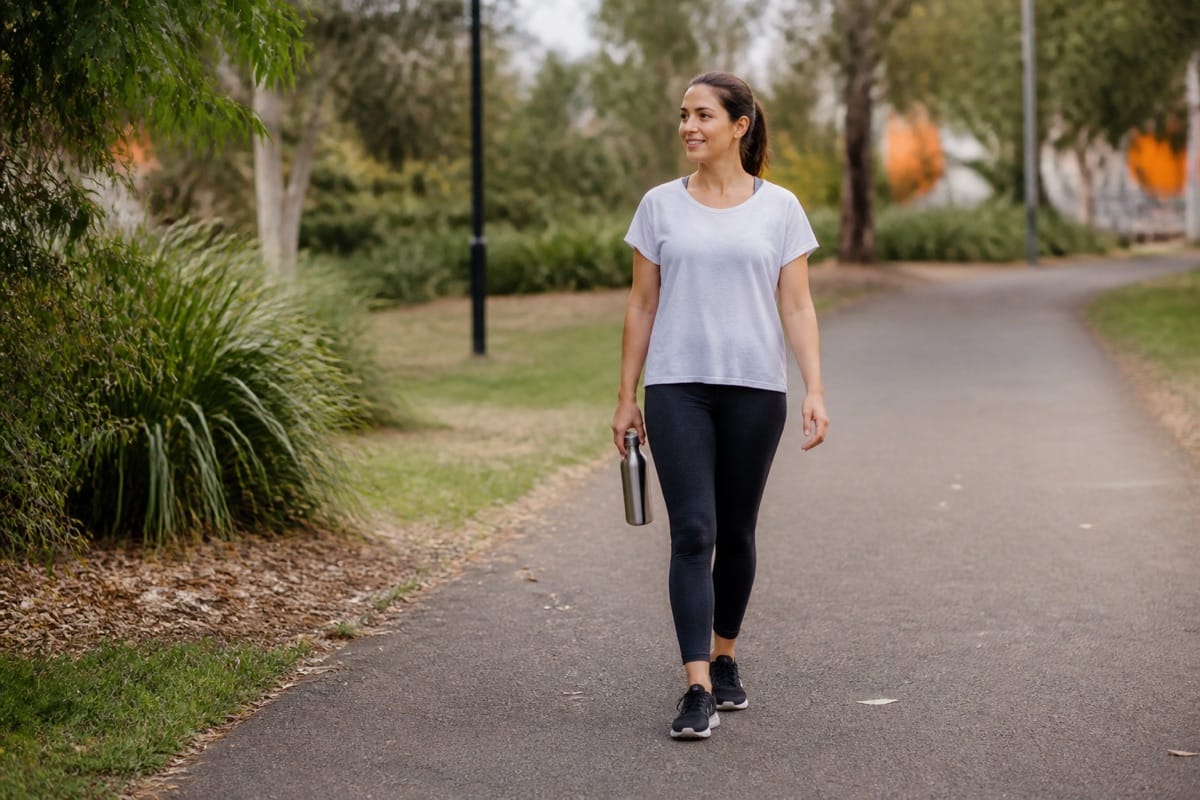 Woman taking a relaxed walk along a leafy park path while carrying a water bottle