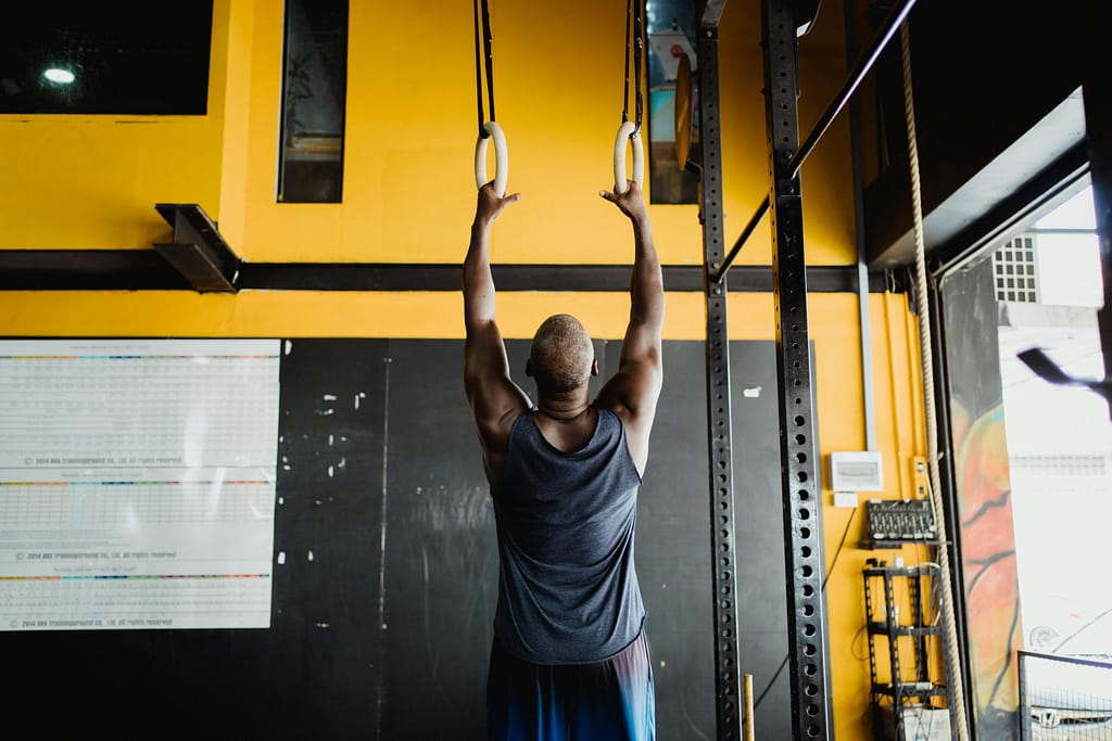 Man hanging from rings. Dead man hang with a yellow background. Great top on fully extending arms up. Shoulder, rotator cuff injury, rehab, exercise, therapy, Preston at Chiropractor Andrew Cunningham Advanced Health
